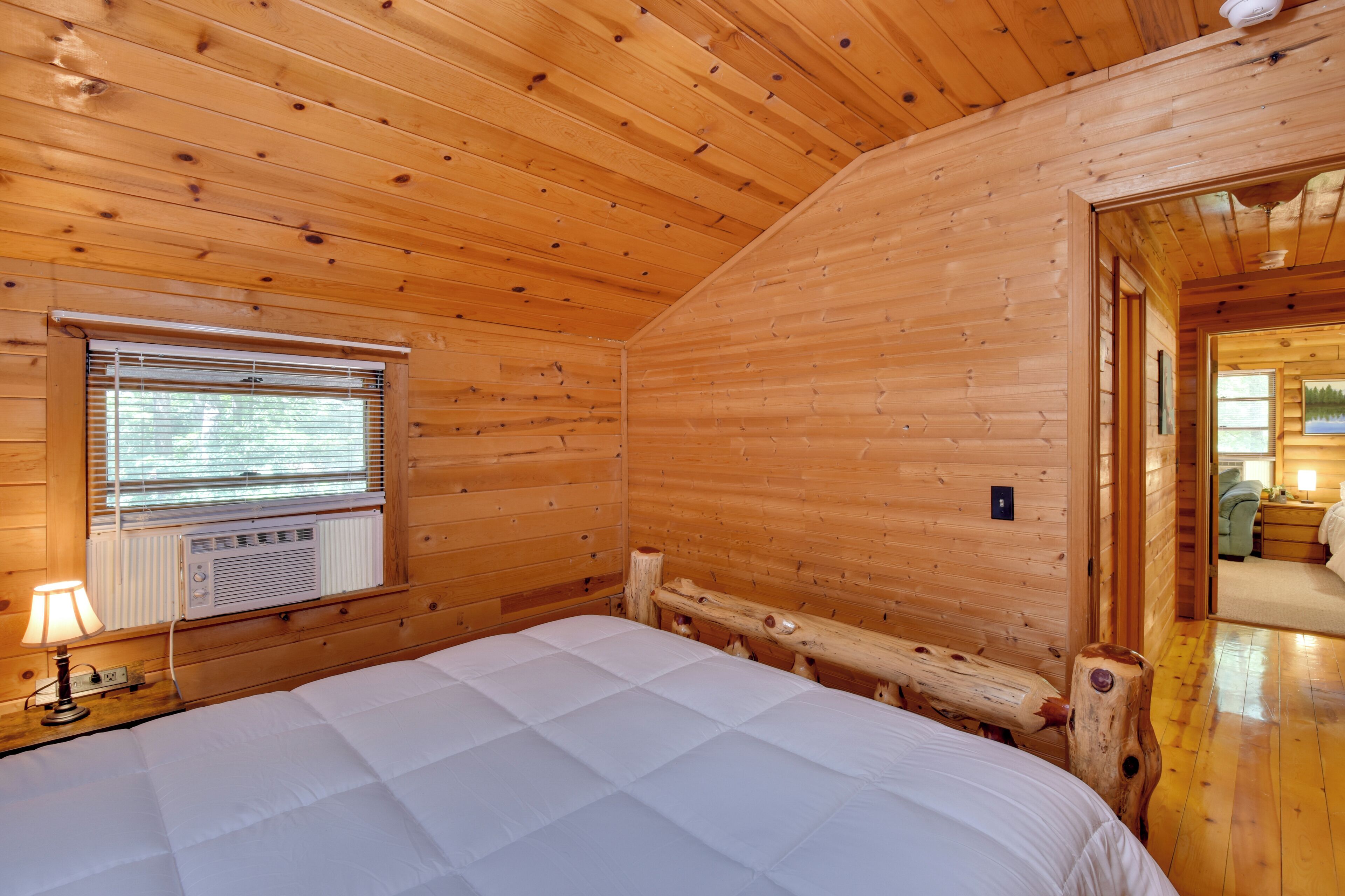A wooden cabin room with a bed, a window with blinds, an air conditioning unit, and a wooden bench.