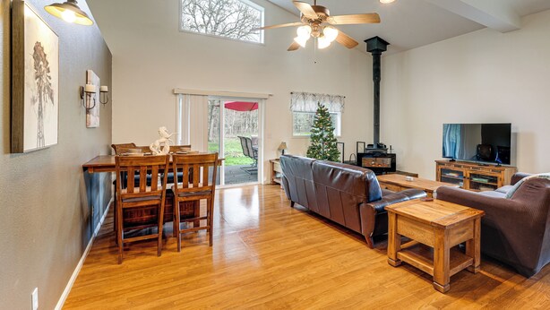 A spacious living room with a wooden floor, a ceiling fan, a fireplace, a television, and a dining area with wooden furniture. - Willamette Valley Home on Working Horse Farm! (Scio)