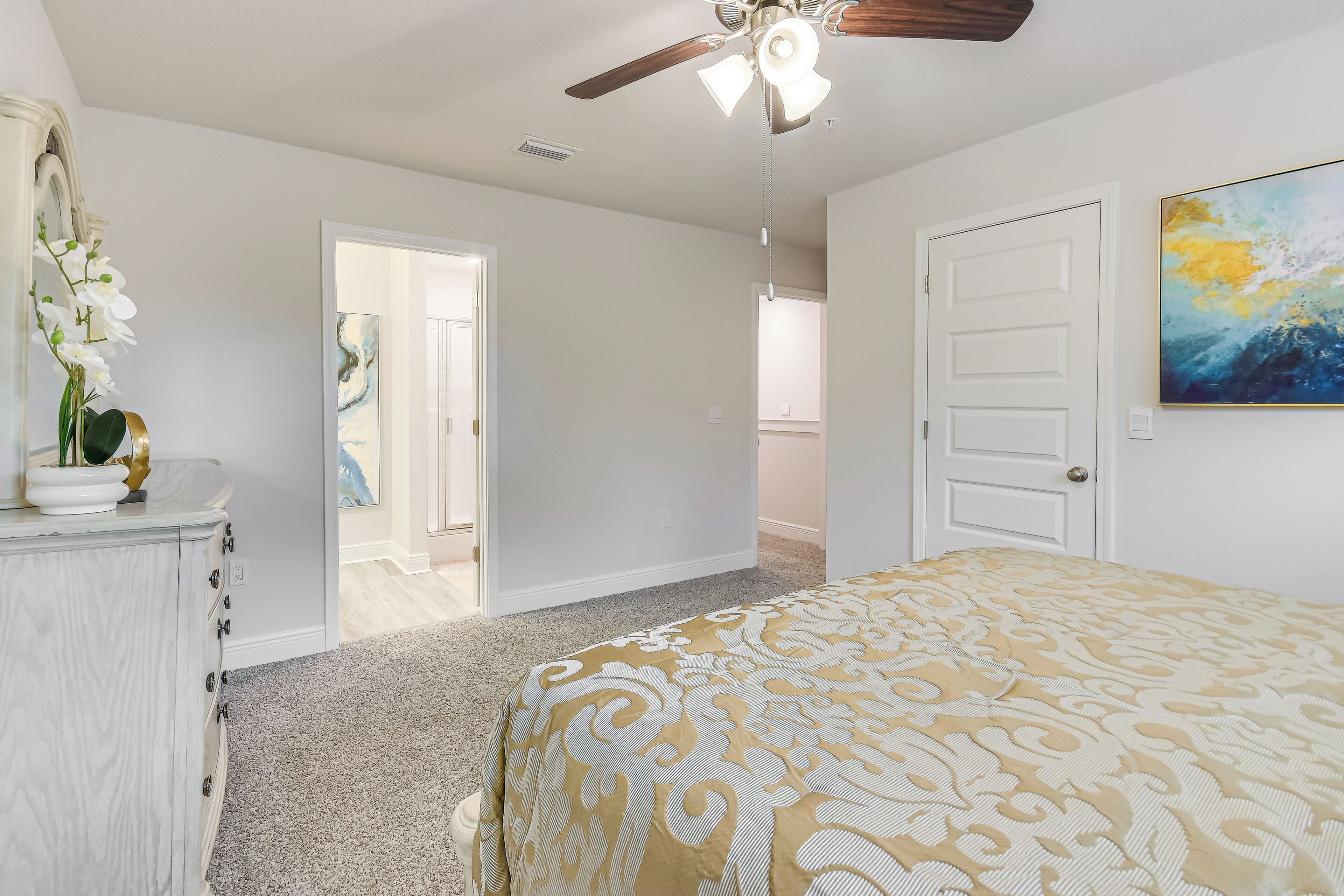 A bedroom with a bed, a dresser, a ceiling fan, and a door leading to another room.