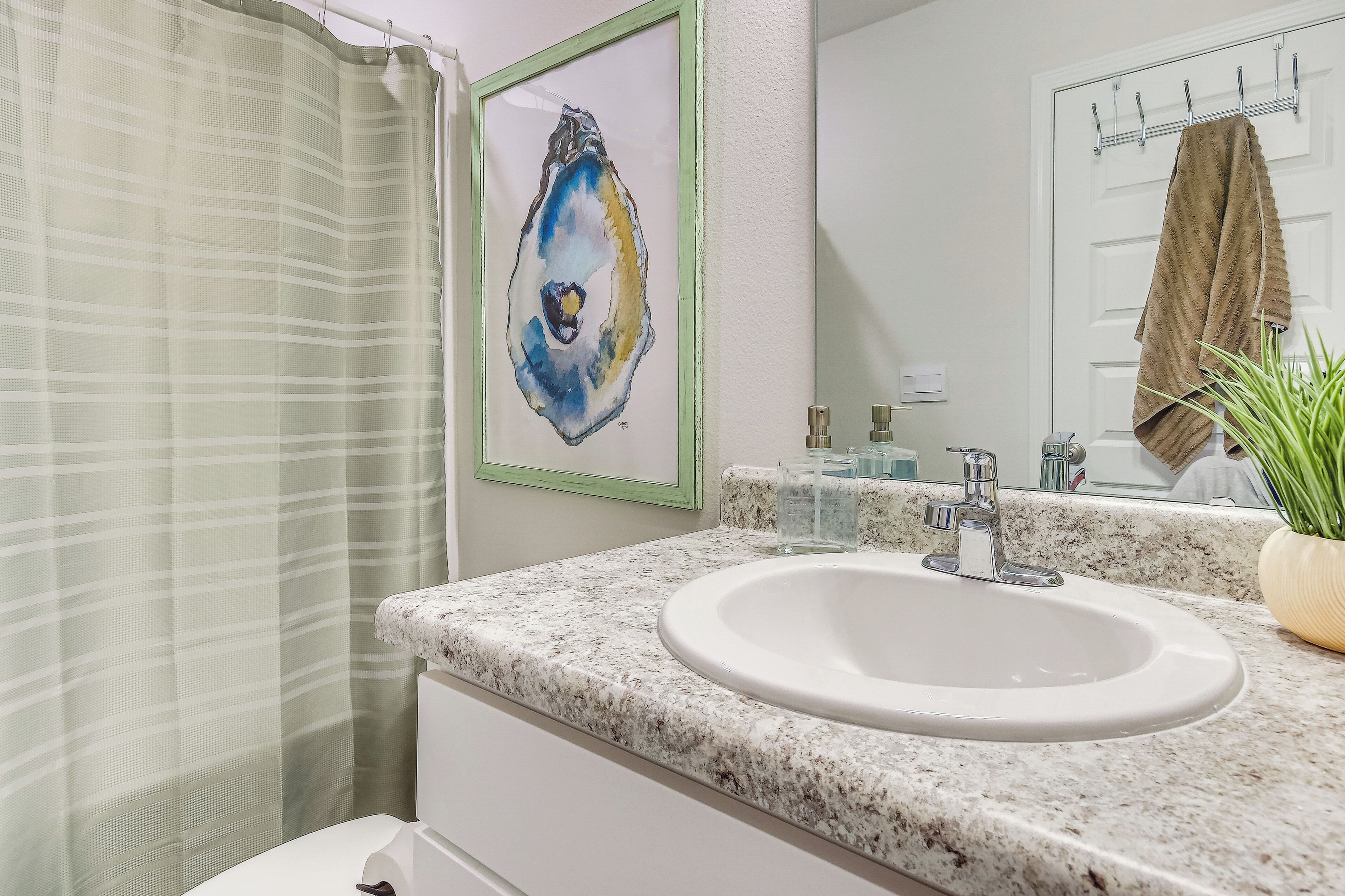 A bathroom with a shower curtain, a sink with a marble countertop, a framed artwork, a towel hanging on a rack, and a potted plant.