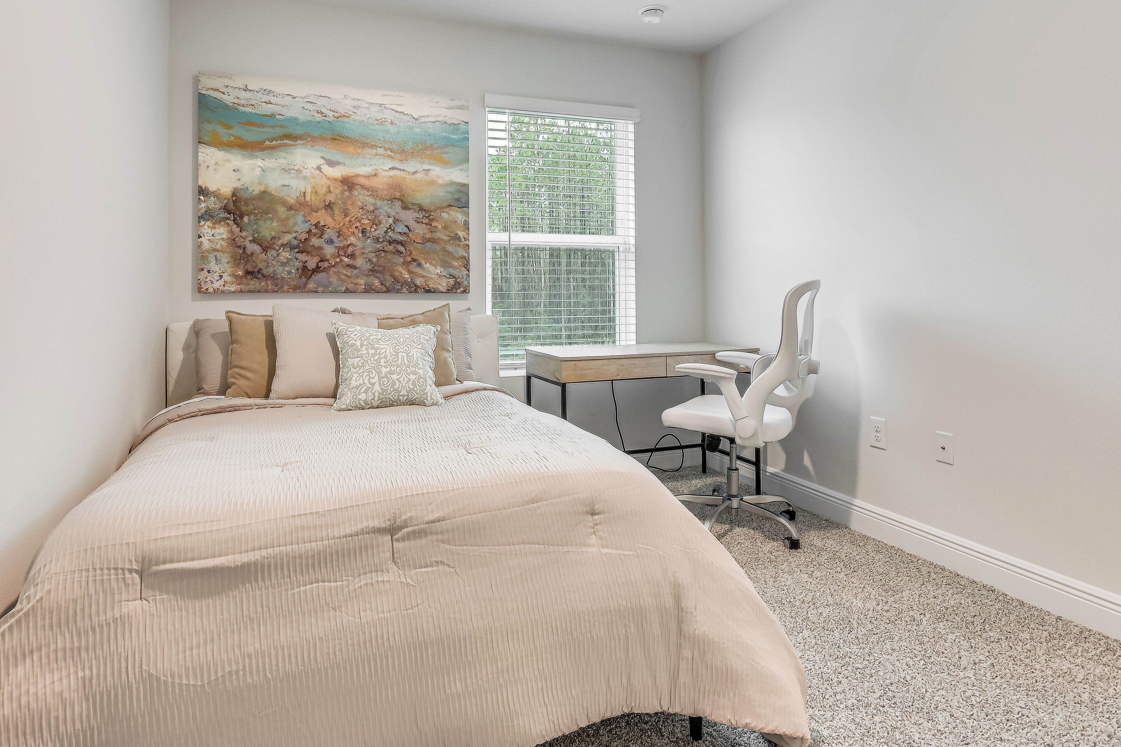A bedroom with a bed, a desk, and a chair. There is a window with blinds.