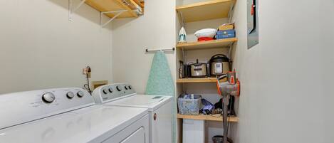 A laundry area with a front-loading washer and dryer, a drying rack, and shelves with various items including a microwave, a kettle, and storage boxes.
