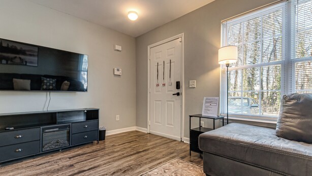 A room with a wooden floor, a grey sofa, a TV mounted on the wall, a door with a window, and a small table with a lamp. - 2 Mi to Lenovo Center: Dog-friendly Getaway (Raleigh)