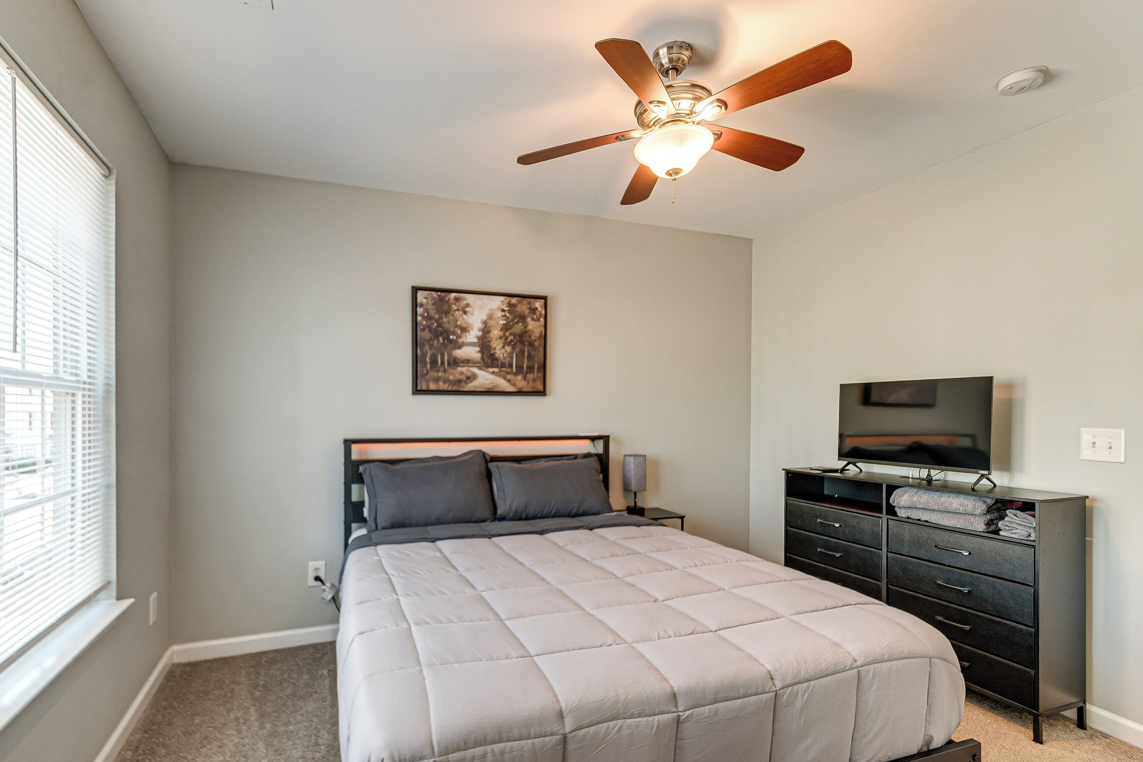 Une chambre à coucher comprenant un lit, un ventilateur de plafond, une commode, un téléviseur et une fenêtre avec des stores.