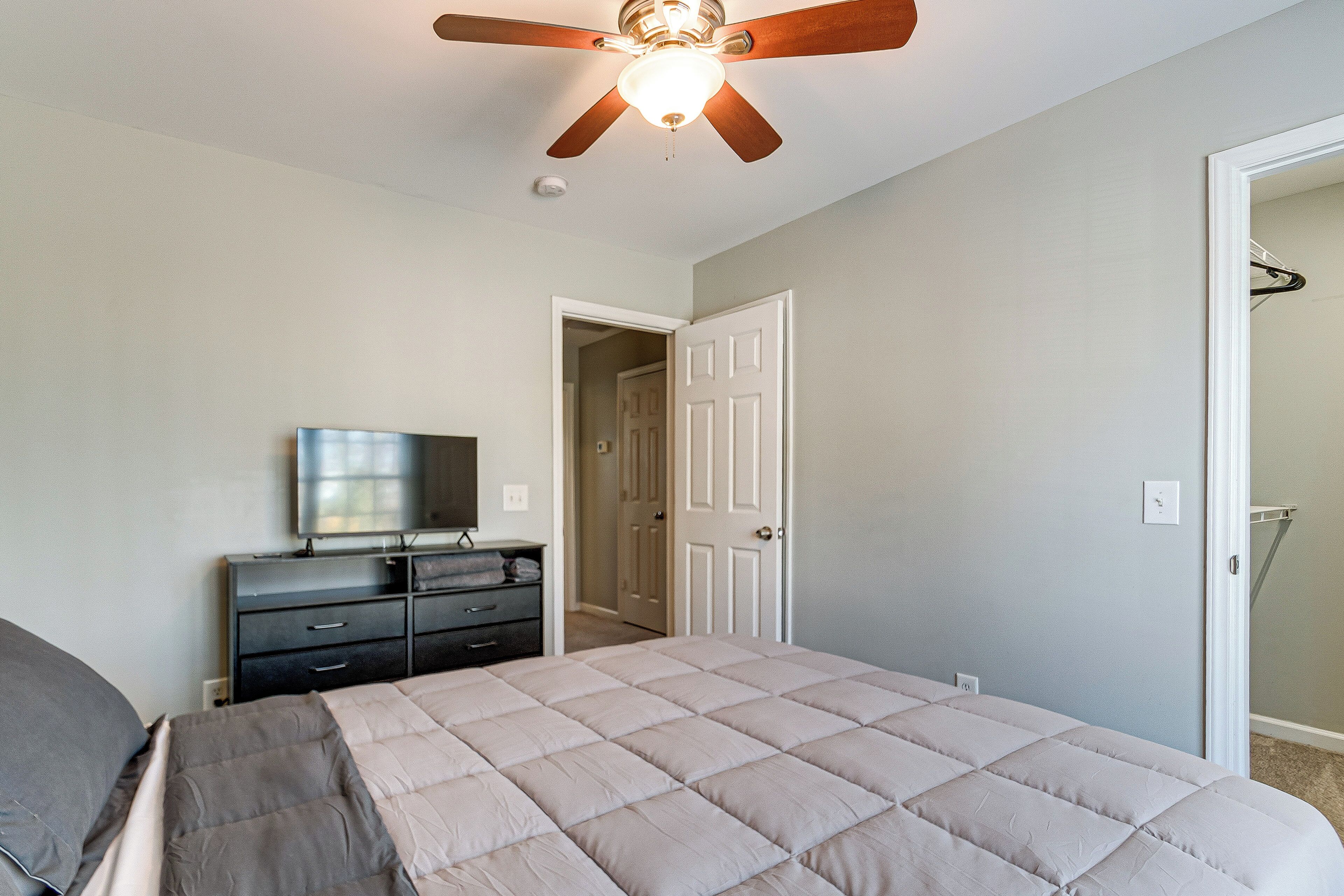 A bedroom with a bed, a dresser, a ceiling fan, and a door leading to another room.