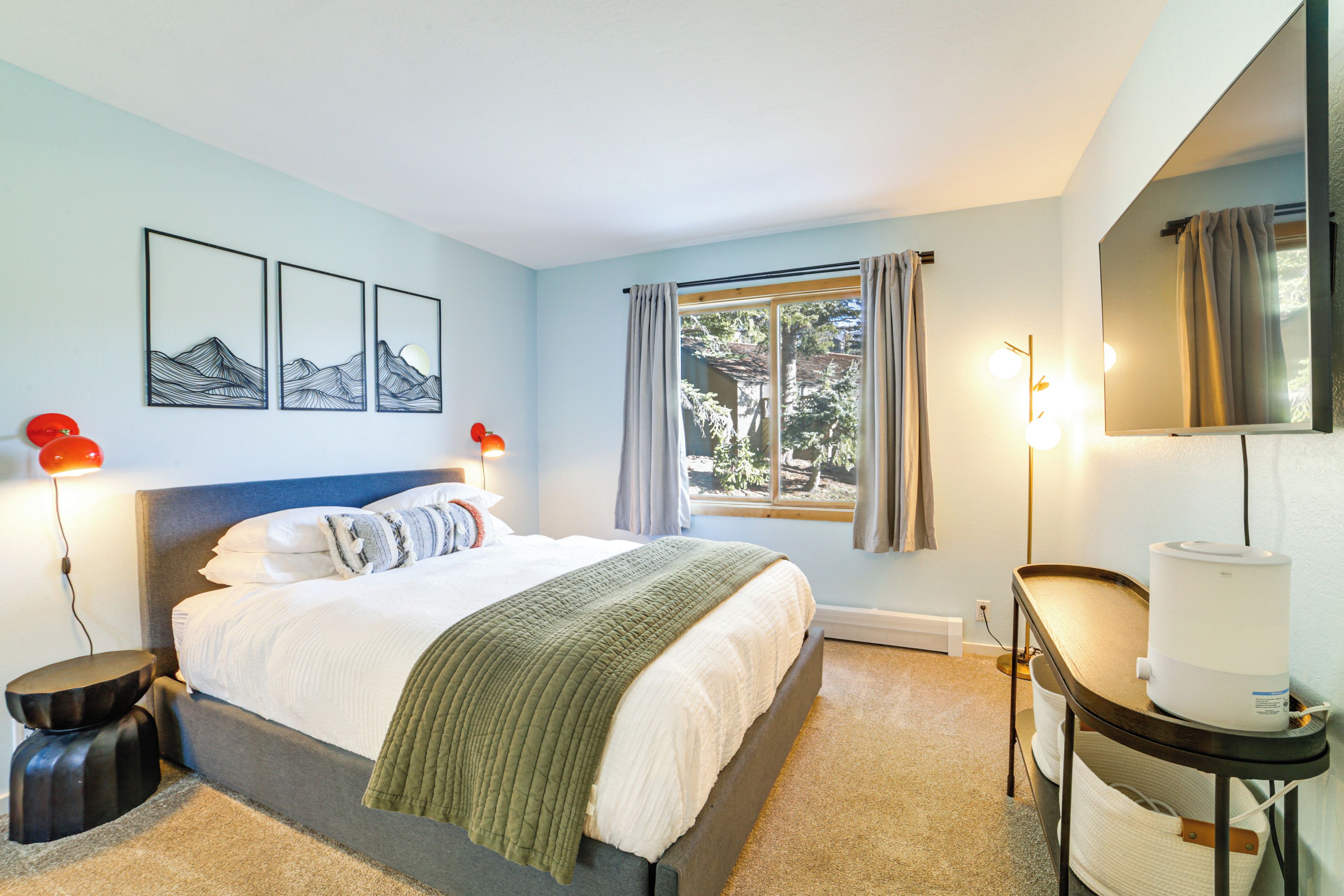 A bedroom with a bed, bedside lamps, a nightstand, a window with curtains, and a wall-mounted mirror.