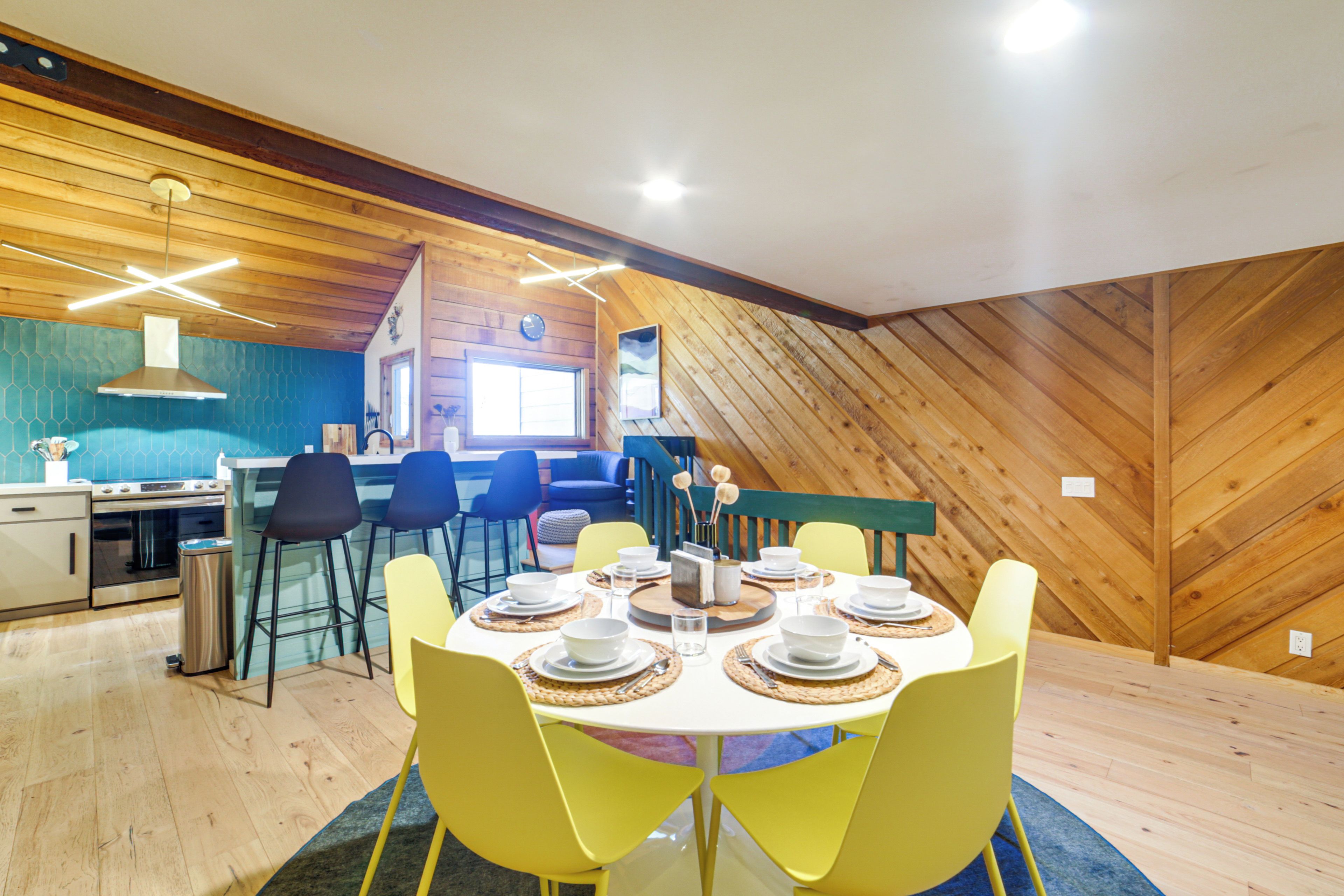 A dining area with a round table set for four, surrounded by yellow chairs. A kitchen with a stove and sink is visible in the background.