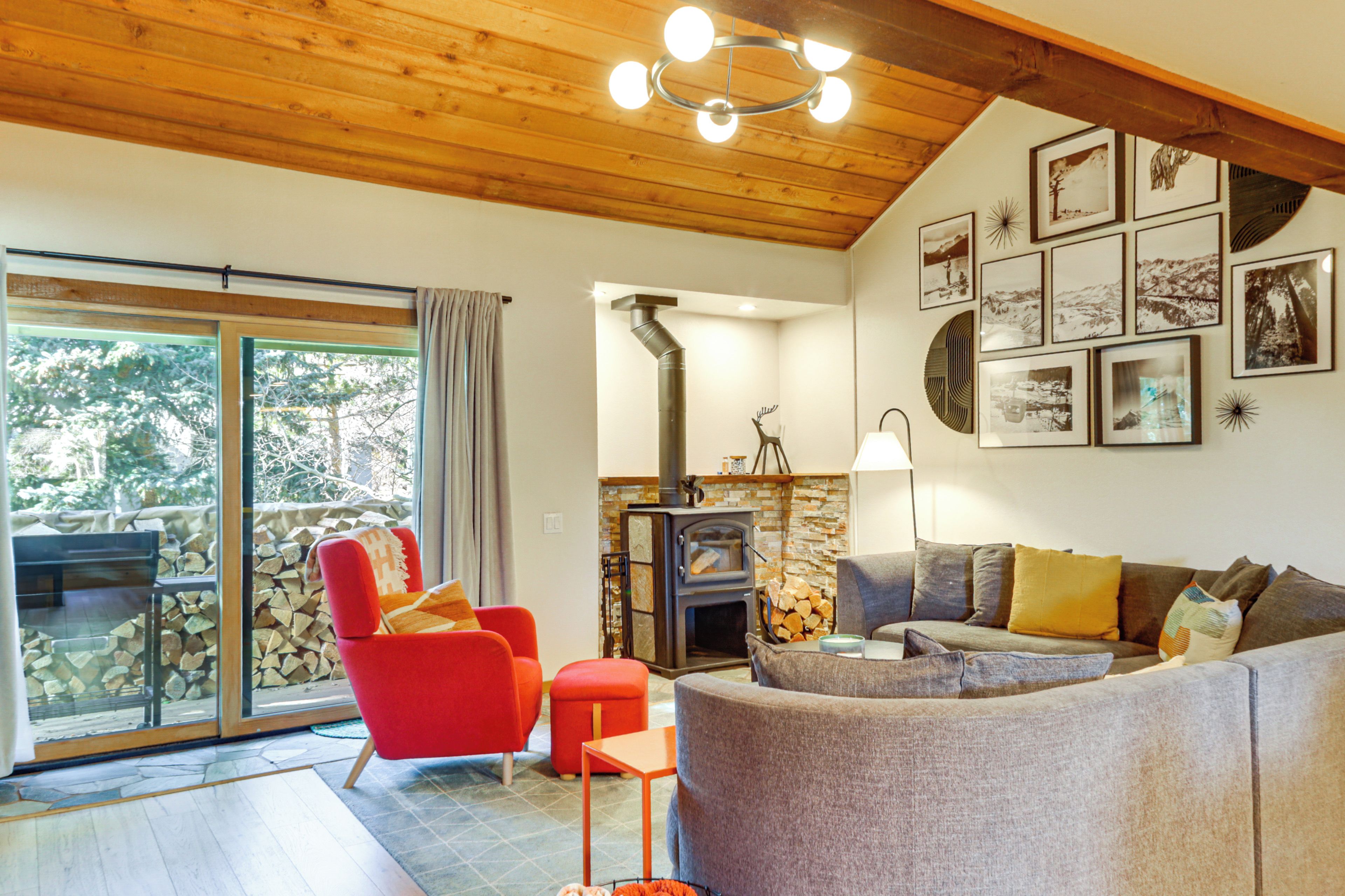 A cozy living room with a fireplace, a red armchair, a grey sofa, and a large window overlooking a forest.