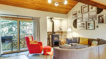 A cozy living room with a fireplace, a red armchair, a grey sofa, and a large window overlooking a forest.