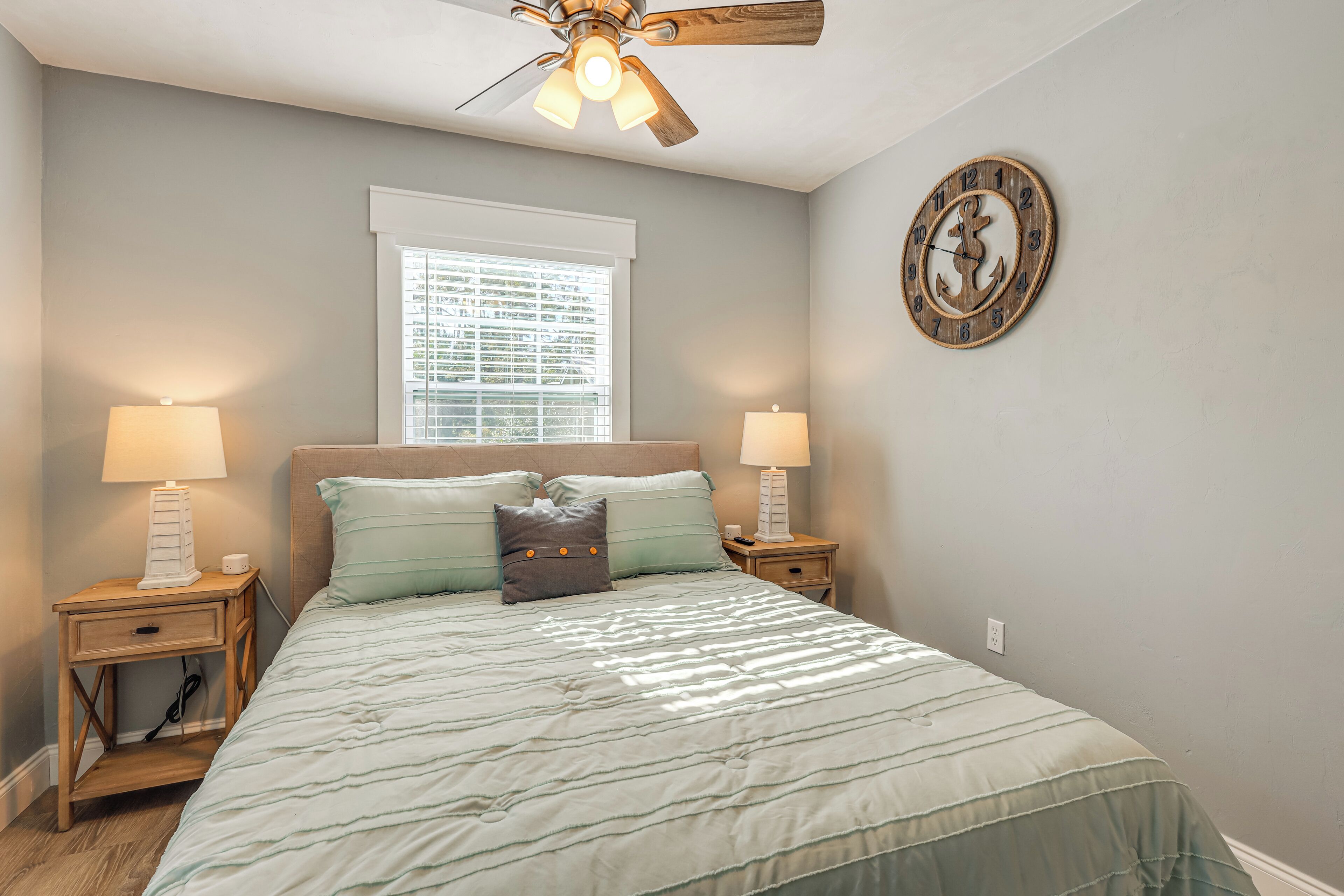 A bedroom with a bed, two bedside tables with lamps, a ceiling fan, a window with blinds, and a wall clock.