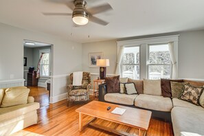 Un salon avec un plancher en bois, un canapé beige, un fauteuil à motifs, une table basse et un ventilateur de plafond.