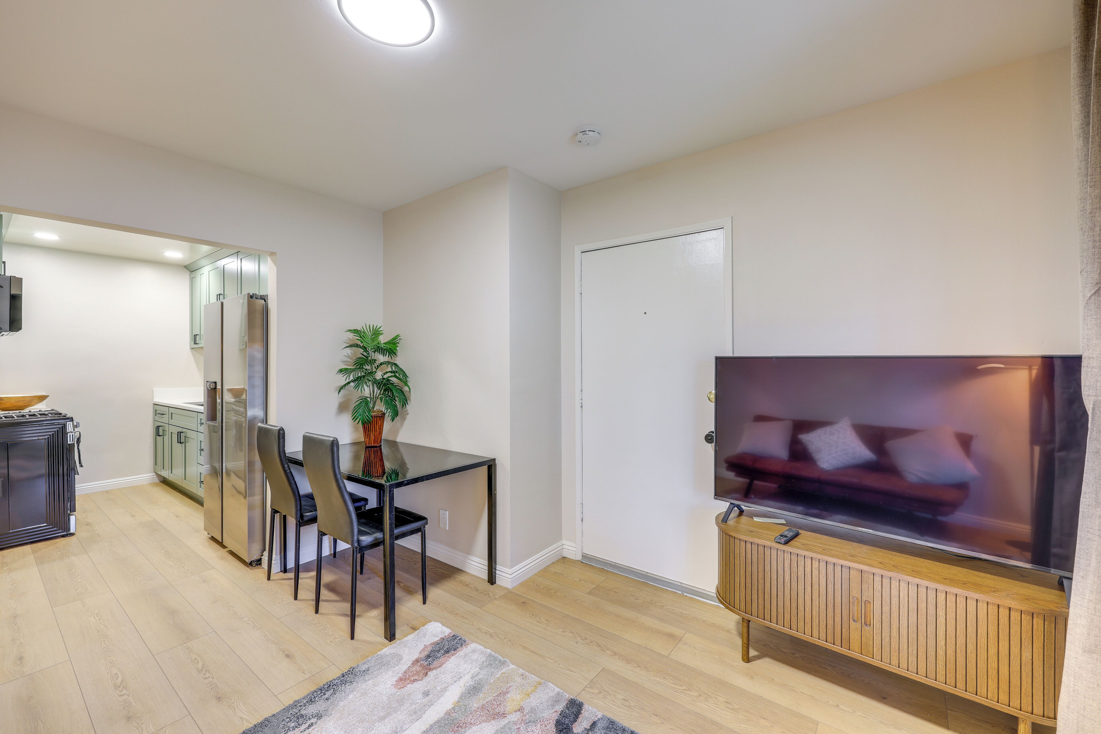 A modern living room with a flat-screen TV, a dining area with chairs and a table, a kitchen with a refrigerator, and a potted plant.