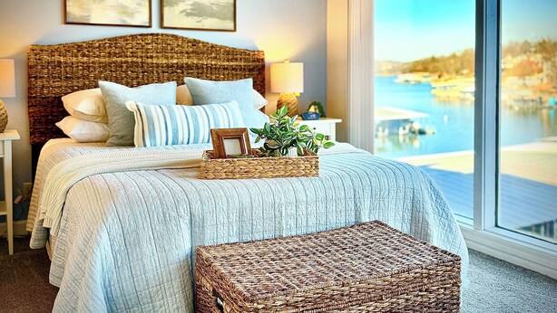 A bedroom with a large bed, a wicker headboard, and a view of a body of water through a large window. - Lake Views, Resort Perks! Osage Beach Retreat (Osage Beach)