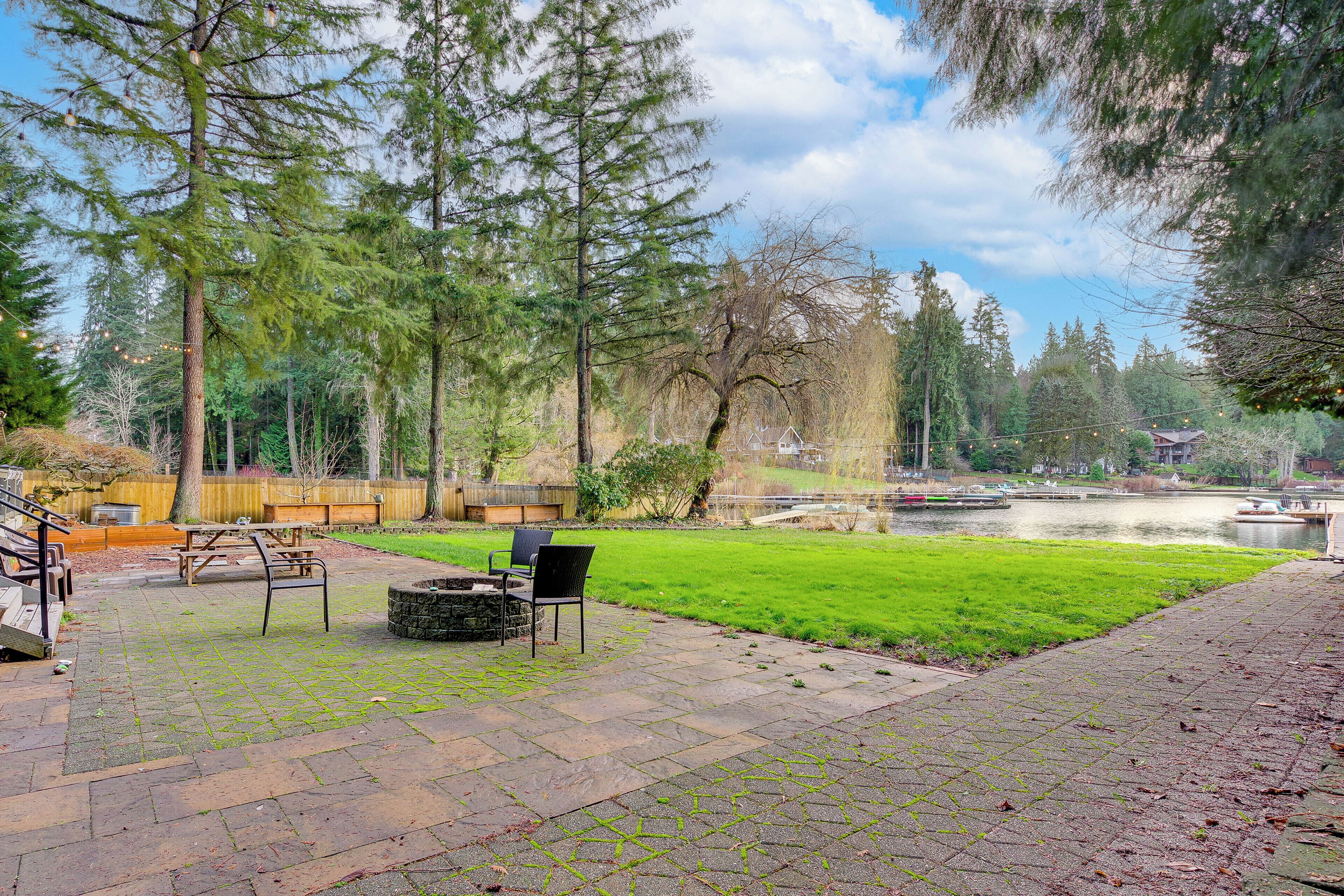 Eine Terrasse mit Picknicktisch und -stühlen, einer Steinkaminfeuerstelle und Blick auf einen See mit Bäumen sowie einem Holzzaun im Hintergrund.