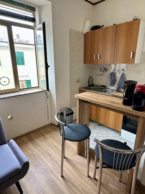 A compact kitchen with wooden cabinets, a sink, and a coffee maker. There is a window with blinds, a chair, and a trash bin. - Cozy apartment in lovely Trieste (Trieste)