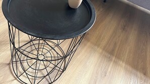 Harmaa sohva, jossa on tyyny nojallaan seinää vasten, pyöreä musta pöytä, jonka jalat ovat metallirunkoa, ja pieni valkoinen kuppi pöydällä.