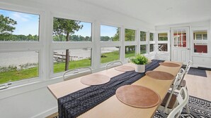 A dining area with a long table, chairs, and large windows overlooking a body of water. - Newly Remodeled Lakeside Cottage. Sleeps 6! (Scottville)