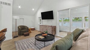 A modern living room with a ceiling fan, a flat-screen TV, a sofa, a coffee table, and a view of a body of water through large windows. - Newly Remodeled Lakeside Cottage. Sleeps 6! (Scottville)