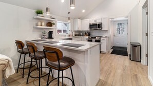 A modern kitchen with white cabinetry, a central island with bar stools, stainless steel appliances, and pendant lighting. - Newly Remodeled Lakeside Cottage. Sleeps 6! (Scottville)