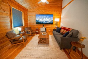 L’intérieur chaleureux d’un chalet en rondins, avec un téléviseur affichant le message « BIENVENUE EN ALASKA » et un décor rustique en bois.