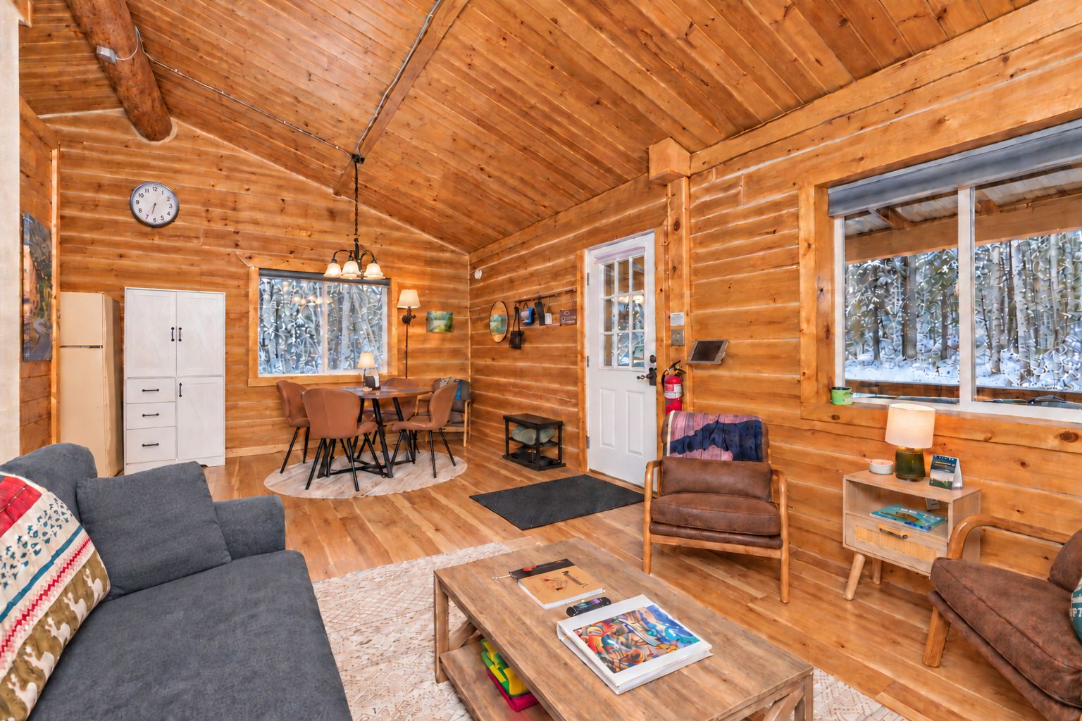 Interior acogedor de una cabaña de madera con comedor, sofá, sillón y chimenea.