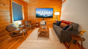 L’intérieur chaleureux d’un chalet en rondins, avec un téléviseur affichant le message « BIENVENUE EN ALASKA » et un décor rustique en bois.