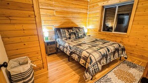 Une chambre confortable dans un chalet, avec un intérieur en bois, un lit recouvert d’une couverture à carreaux, une table de chevet avec une lampe et une fenêtre donnant sur un paysage agréable.