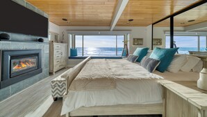 Een slaapkamer met een open haard, een bed met veel kussens, uitzicht op de oceaan en een houten plafond.