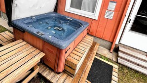 Un bain à remous doté de bancs en bois et d’une terrasse en bois, situé à côté d’un bâtiment rouge avec des fenêtres et une porte.