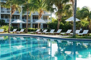 A pool area with lounge chairs and umbrellas, surrounded by palm trees and a building with balconies. - Top Floor, Oceanfront 1-bedroom suite w/balcony and the best view of Grace Bay (Grace Bay)