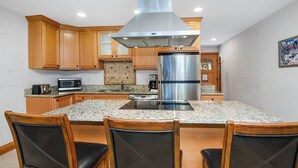 Una cocina con armarios de madera, una campana extractora de acero inoxidable, un refrigerador de doble puerta, encimeras de granito y tres sillas de madera con cojines negros.