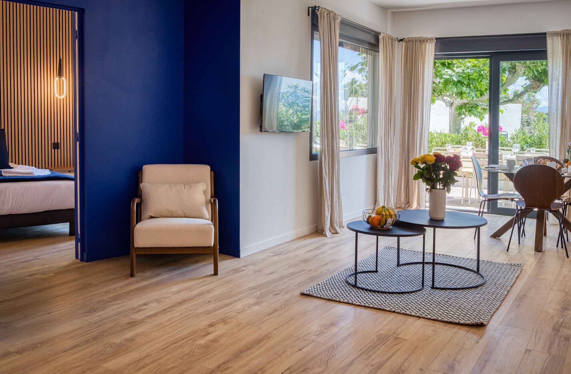 Una habitación moderna con cama, una silla, una mesa con un jarrón de flores y vistas al exterior a través de amplias ventanas.