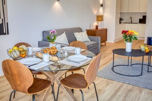 A modern dining area with a glass table, wooden chairs, and a grey sofa in the background. - UrbanFlat 213 - Aura Plage 02 - Sunrise (Roquebrune-sur-Argens)