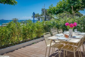An outdoor dining area with a view of the sea, a wooden deck, and a table set for two with chairs. - UrbanFlat 213 - Aura Plage 02 - Sunrise (Roquebrune-sur-Argens)