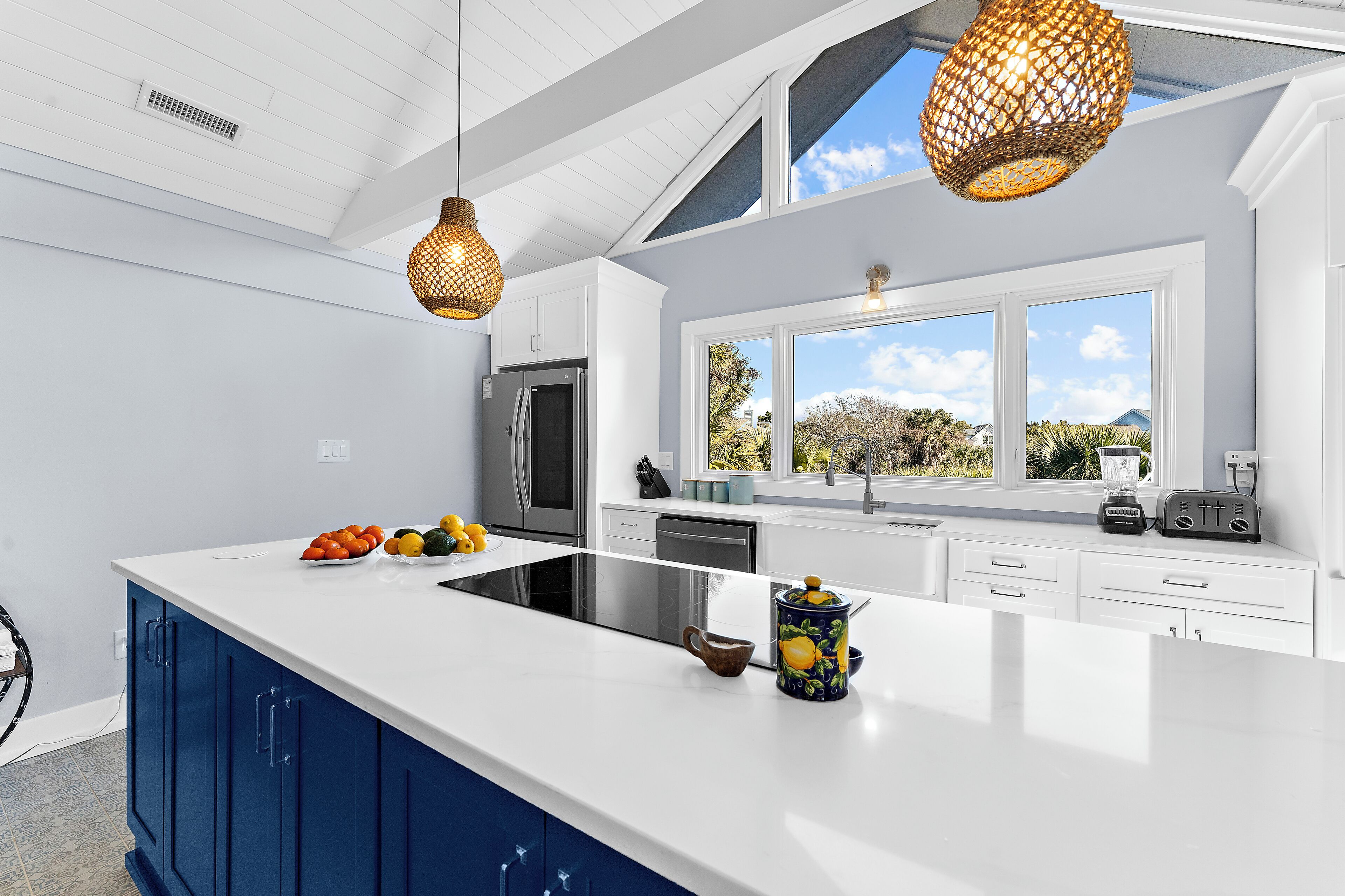 A modern kitchen with a large island, white countertops, blue cabinets, and a view of the outdoors through a large window.