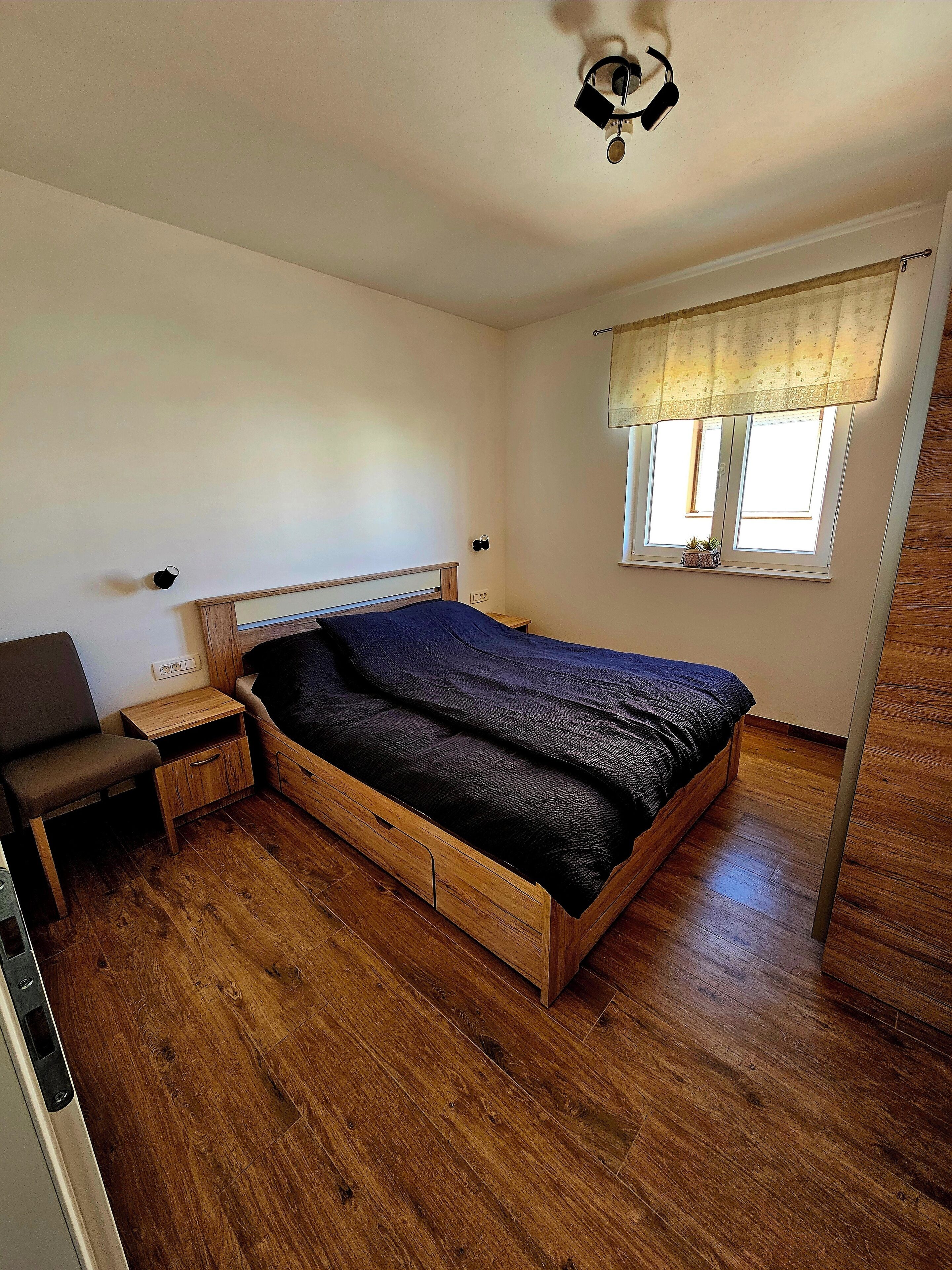 A bedroom with a wooden bed, a nightstand, a chair, a window with a curtain, and a ceiling fan.