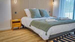 Ein ordentlich bezogenes Bett mit einer grün-weißen Decke, zwei Kissen, einem Nachttisch aus Holz mit einer Lampe und einem Fenster mit hellblauen Vorhängen.