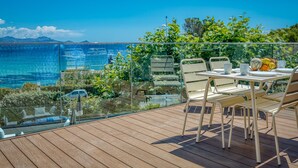 Eine Holzterrasse mit Sitzgelegenheiten im Freien, einem Glasgeländer und Blick auf das Meer und die Berge.