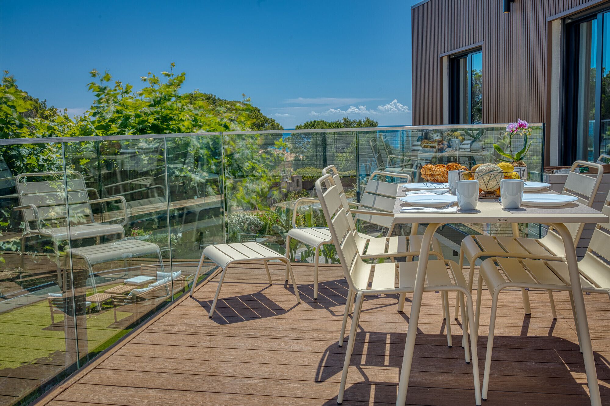 Une terrasse avec des sièges et une table dressée pour les repas, offrant une vue sur un jardin luxuriant et un bâtiment moderne.