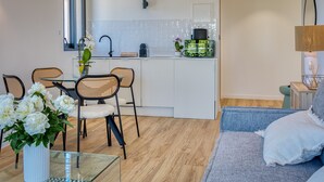 A modern kitchen with white cabinets, a black faucet, and a small island with a green appliance. There is a dining area with a glass table and wicker chairs, and a living area with a blue sofa and a lamp. - UrbanFlat 219 - Aura Plage 08 - Aquamoon (Roquebrune-sur-Argens)