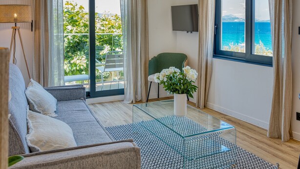 A modern living room with a sofa, a glass coffee table, and a chair. There is a TV mounted on the wall and a balcony with a view of the sea. - UrbanFlat 219 - Aura Plage 08 - Aquamoon (Roquebrune-sur-Argens)