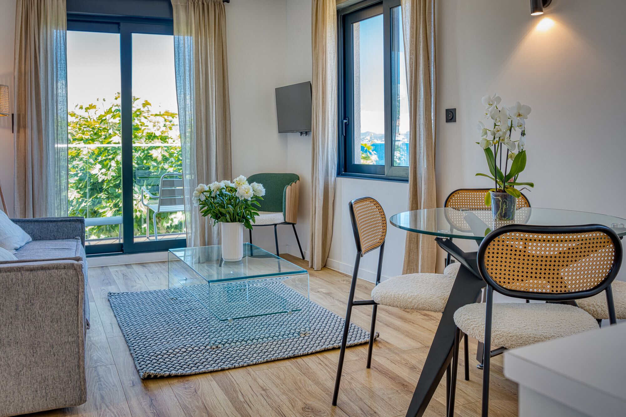 A modern living room with a glass dining table, wicker chairs, a sofa, and a view of greenery outside.