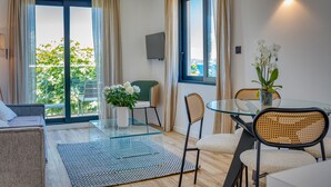 A modern living room with a glass dining table, wicker chairs, a sofa, and a view of greenery outside. - UrbanFlat 219 - Aura Plage 08 - Aquamoon (Roquebrune-sur-Argens)