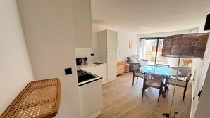 Una cocina moderna con armarios blancos, un comedor con mesa de vidrio y sillas de mimbre, y un pequeño balcón con mesa de mimbre.