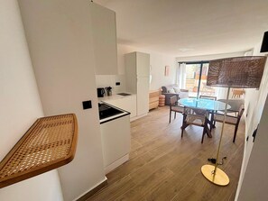 A modern kitchen with white cabinets, a dining area with a glass table and wicker chairs, and a small balcony with a wicker table. - UrbanFlat 221 - Aura Plage 10 - Sea Sun (Roquebrune-sur-Argens)