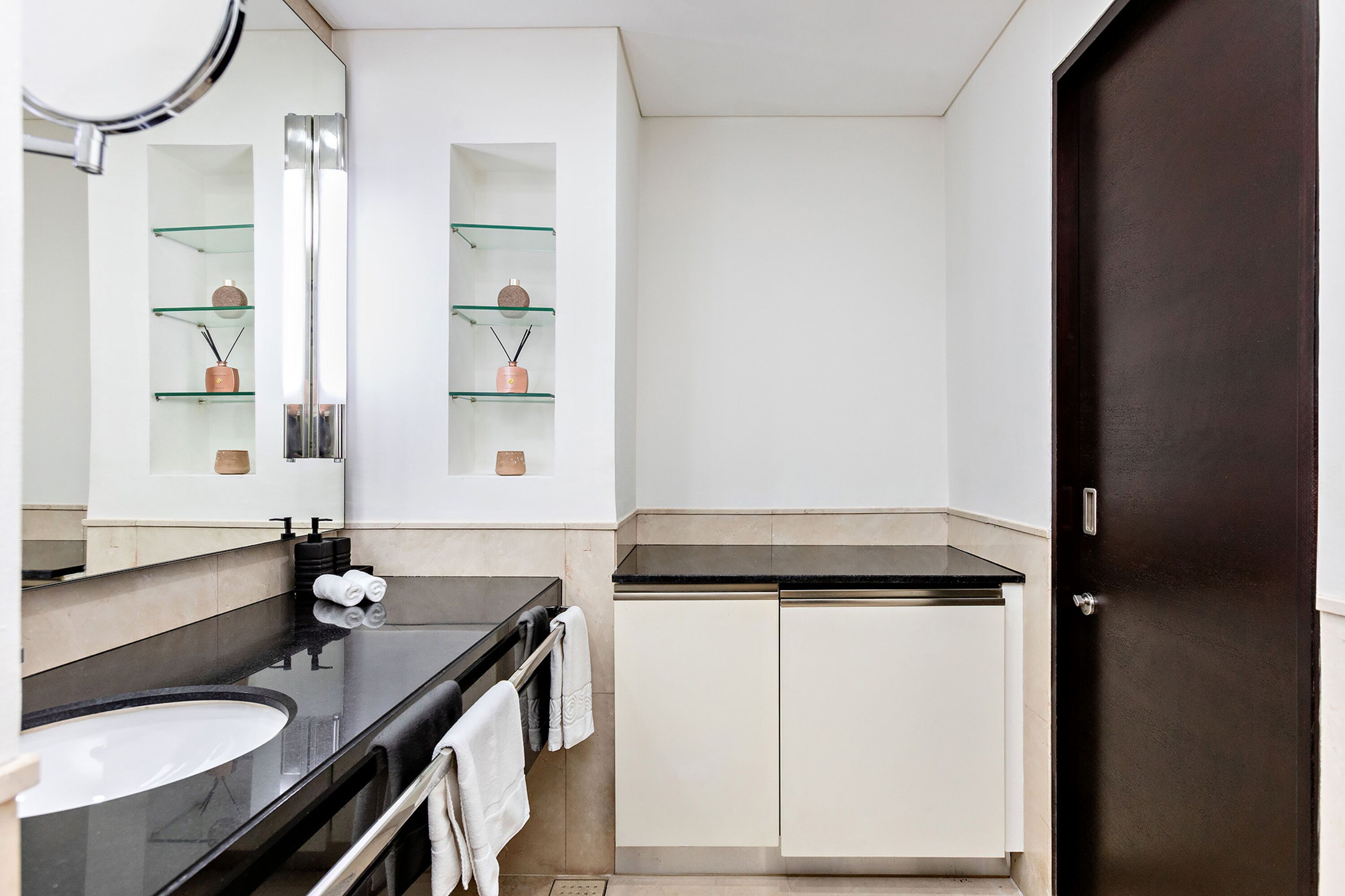 Une salle de bain moderne avec un comptoir noir, des armoires blanches, un grand miroir et des étagères en verre.