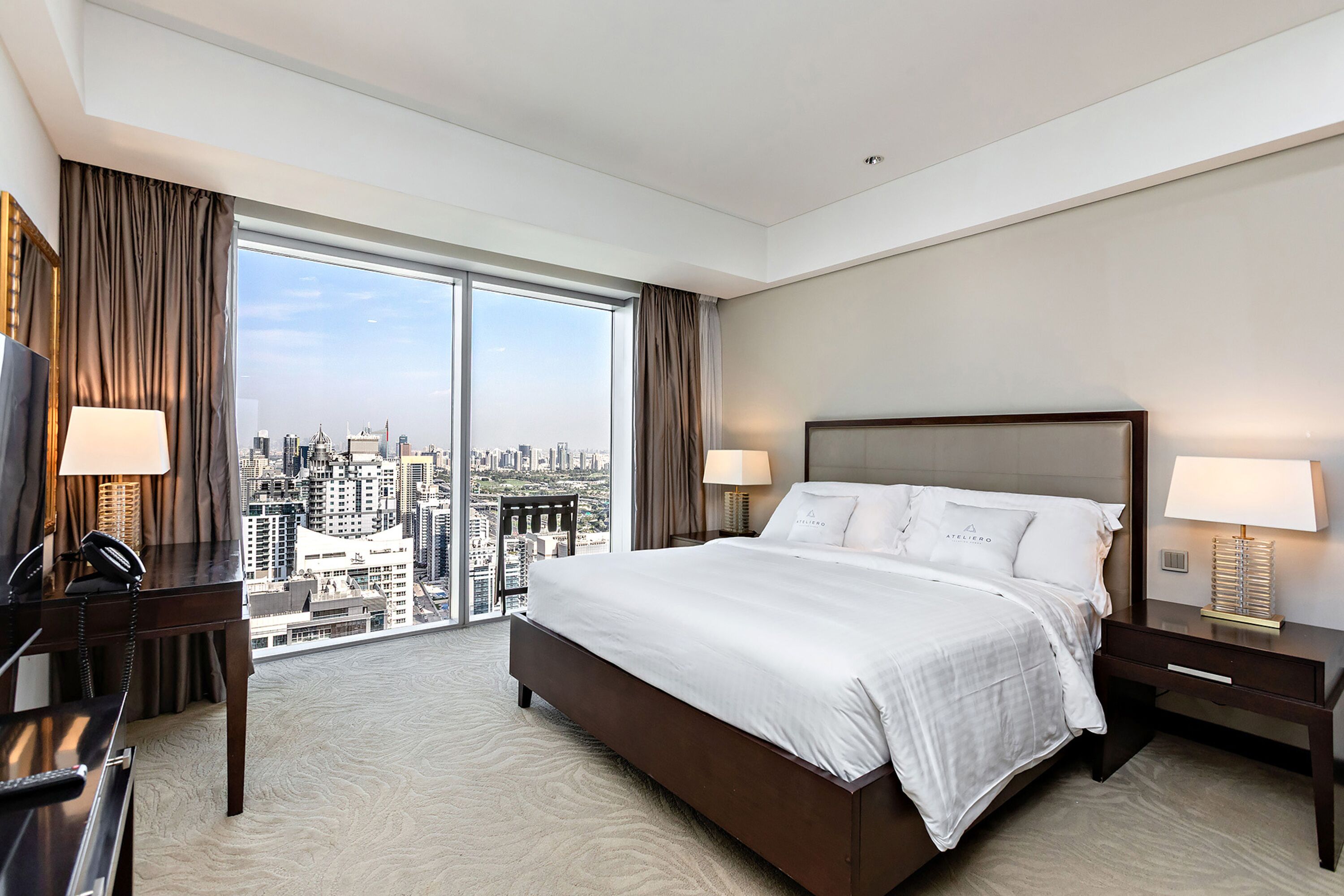 A hotel room with a large bed, two bedside tables with lamps, a desk with a telephone, and a view of a cityscape through large windows.