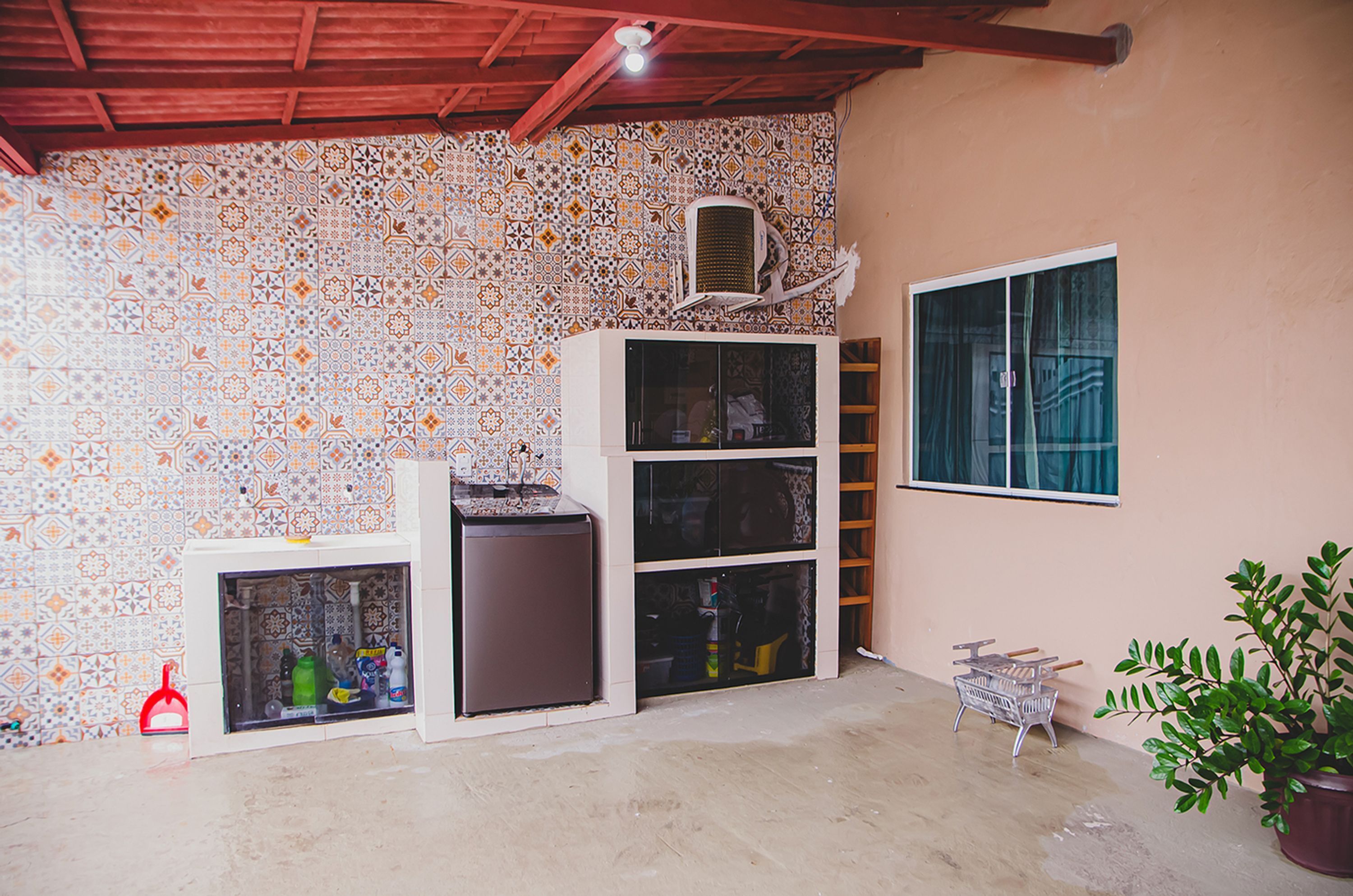 A room with patterned tile walls, a white cabinet with glass doors, a small refrigerator, a window with curtains, a ladder, a plant, and a metal basket.