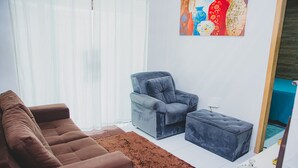 A living room with a brown sofa, a grey armchair, and a matching ottoman. There is a painting on the wall and a window with sheer curtains. - Casa Pet Friendly em Condom nio c Vaga em Marituba (Marituba)