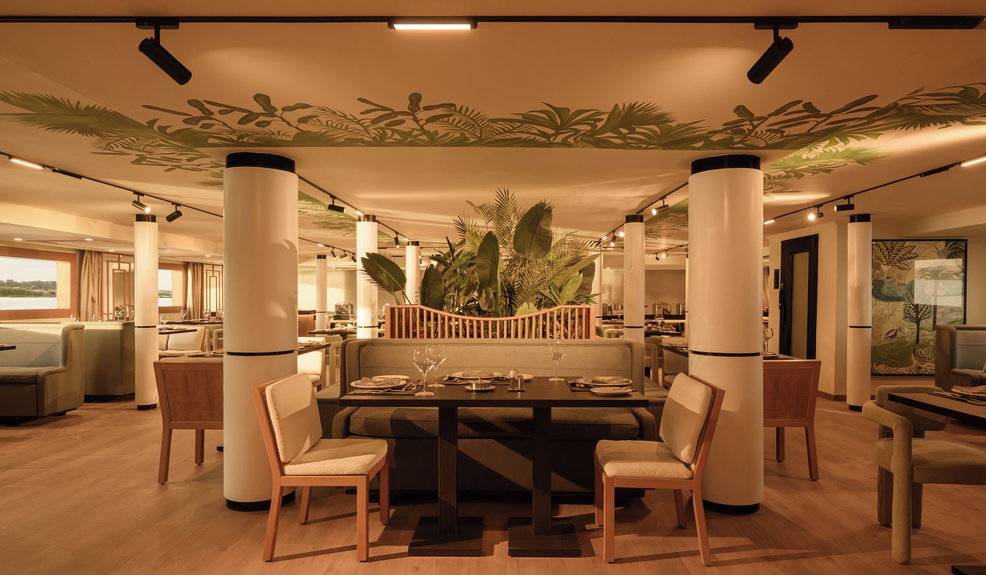A dining area with a central table, surrounded by chairs, and a decorative plant barrier.